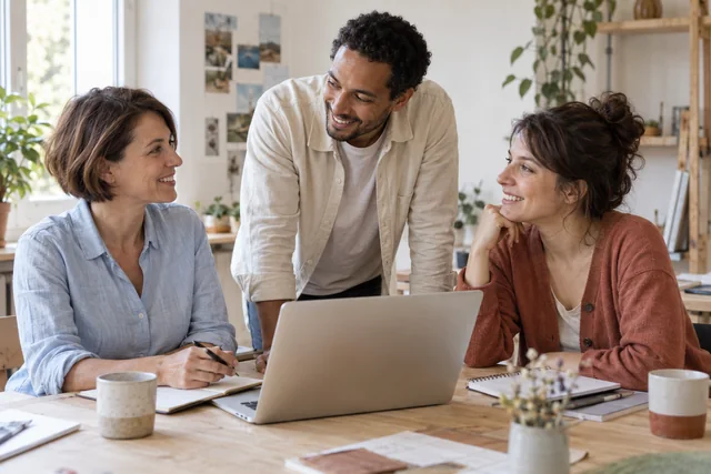 Trois collègues échangent avec le sourire autour d’une table de travail.